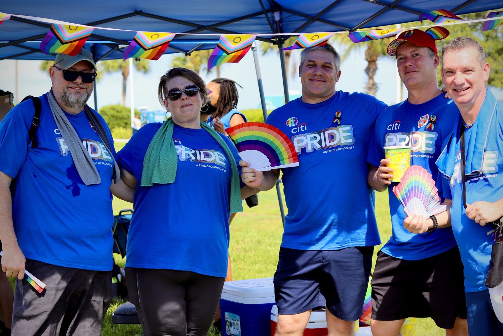 Five team members in blue Pride shirts posing together under a tent with rainbow fans