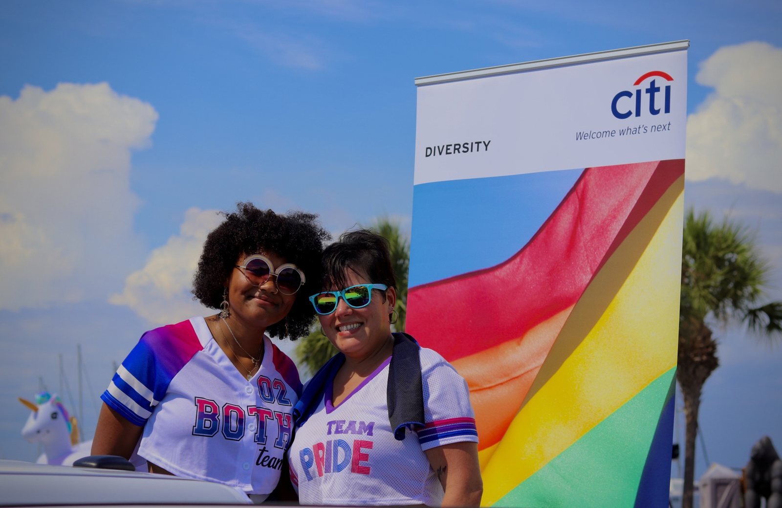 Two marchers smiling in front of a rainbow banner and palm trees